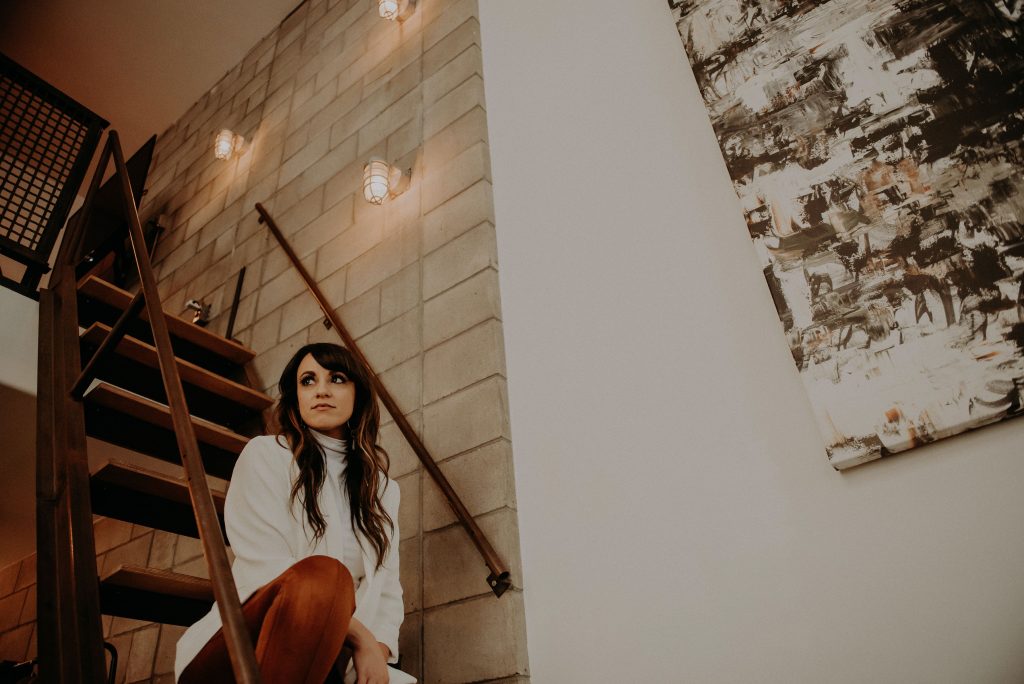 brand designer in white blazer and rust pants sitting on stairs in creative space shares how to niche down your design business